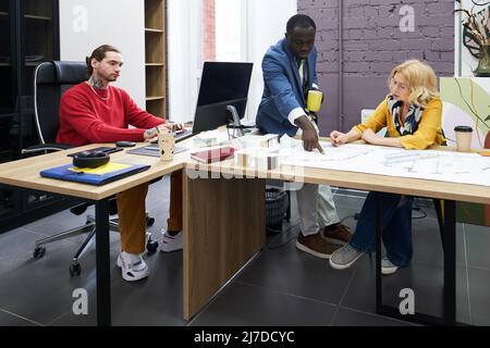 Two architects sitting at table and working over blueprint together with their colleague using computer at his workplace Stock Photo
