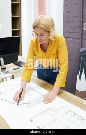 Serious female engineer in eyeglasses working with blueprint on table using pencil and ruler Stock Photo