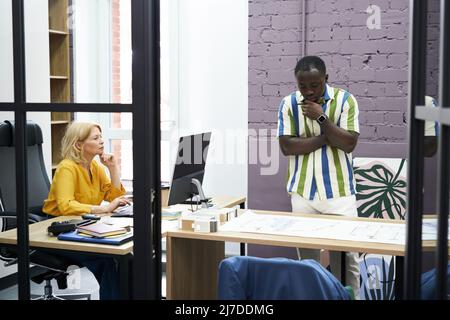 African businessman examining blueprint on table with businesswoman working at her workplace on computer during work at office Stock Photo