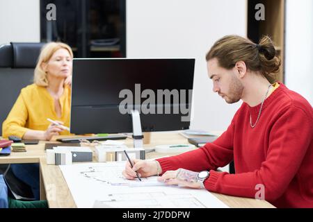 Young professional designer drawing design building project in office with his colleague working on computer in background Stock Photo