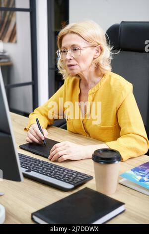 Serious mature woman in eyeglasses sitting at her workplace with computer using graphic tablet at her work Stock Photo