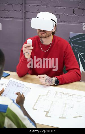Young designer sitting at table with blueprint and using virtual reality headset to look at new project Stock Photo