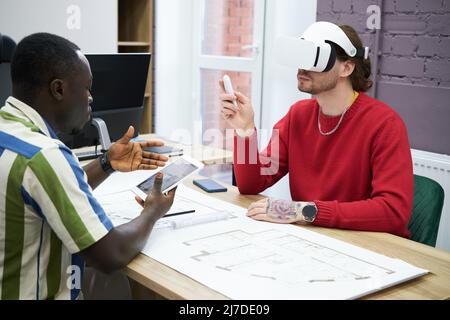 Business people sitting at table with blueprint using digital tablet to connect with VR goggles during teamwork at office Stock Photo