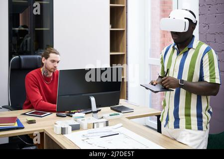 Graphic designer using VR headset to draw blueprint with tablet with his colleague using computer at table Stock Photo