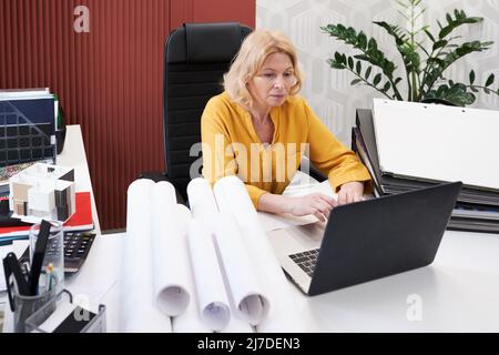Serious mature architect sitting at table with folders and blueprints and typing on laptop, she working over new project at office Stock Photo