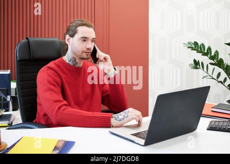 Young businessman sitting at office desk with laptop and having conversation on mobile phone Stock Photo