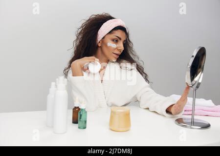 Lovely tanned pretty curly Latin lady admiring herself in mirror use facial cream doing beauty procedure in white home interior. Copy space. MIRROR Stock Photo