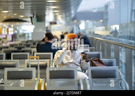 Smiling black traveler man drinking coffee waiting for flying at airport talking on mobile phone Stock Photo