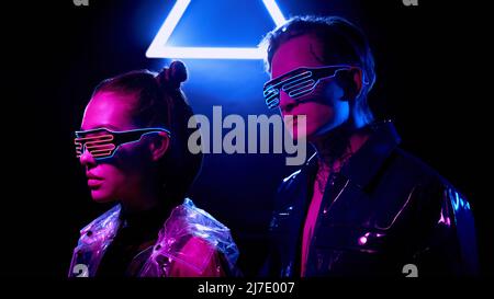 Young cyber couple in vinyl jackets and LED goggles standing in dark room with neon triangle sign Stock Photo