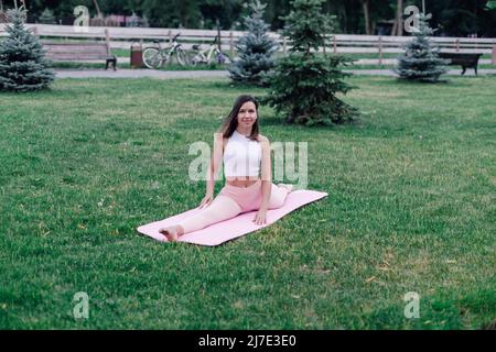 Attractive girl practicing yoga. Brunette in sports clothes is sitting in pose of Monkey god, doing exercises on pink yoga mat in park Stock Photo