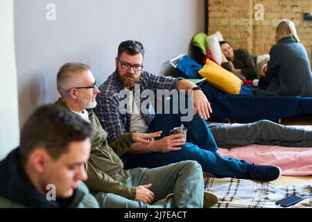 Mature and young refugee men having talk by cup of tea while sitting by wall on mattresses serving them as beds against restful women Stock Photo