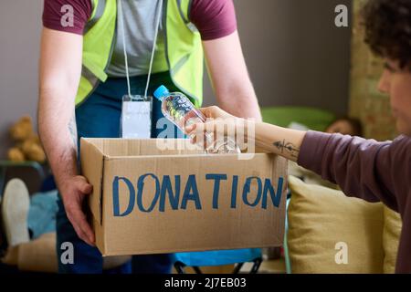 Young male volunteer in uniform holding donation box with food and drinks while female refugee taking bottle of water Stock Photo