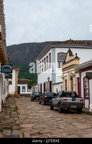 Many colonial buildings at the west end of Direita cobblestone street with cars parked at it in Tiradentes historical center under clouded sky. Stock Photo