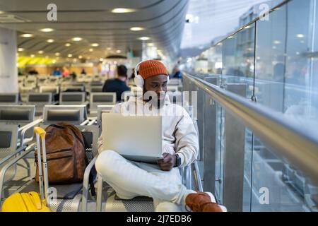 Young hipster black man sitting in airport terminal using laptop wear headphones and listening music Stock Photo