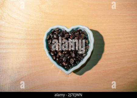 Overhead shot of coffee beans inside a heart shaped white bowl on a wooden textured table - stock photography Stock Photo