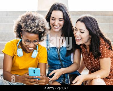 Three multiracial women friends laughing together while using mobile phone Stock Photo