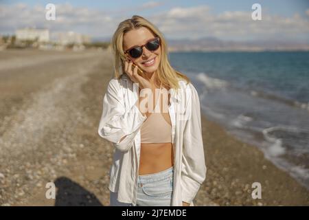 Lovely lady in sunglasses smiling while talking on the phone while standing on a beach Stock Photo