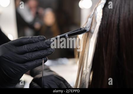 close-up side view of a strand of female hair on a special sheet of foil, the master applies hair dye with a brush. The process of highlighting by a Stock Photo