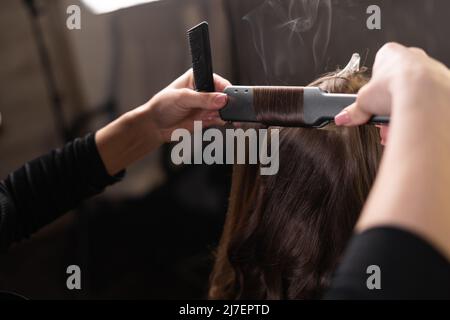 The stylist straightens female hair with an iron in the salon close-up of the master's hand. Stock Photo