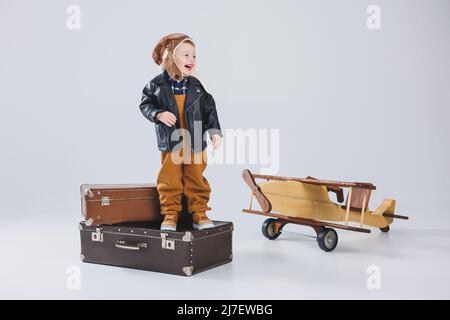 The boy is crying in a leather jacket and a pilot's hat, a wooden plane, brown suitcases. Children's wooden toys. Baby emotions Stock Photo