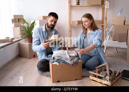 a young couple in love in a new apartment on day of the move sit on the floor unpack things a man holds a hat in his hands. The couple moves into a Stock Photo