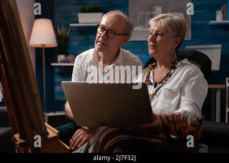 Senior couple sitting in front of easel holding laptop looking at online tutorials to improve drawing technique. Retired man and woman looking for inspiration on social media using portable computer. Stock Photo