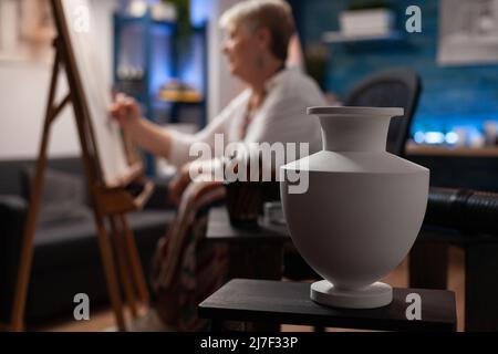 Closeup of white vase on wooden table used for inspiration by retired artist drawing in pencil on easel in home art studio. Selective focus on still life object used as subject for sketch in workshop. Stock Photo