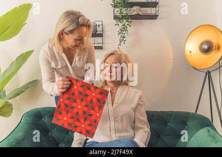 Smiling grown up daughter congratulating happy mature mother and giving birthday gift, excited senior grandma receiving surprise on Mothers Day from adult granddaughter. High quality photo Stock Photo