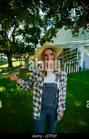 Caucasian smiling female farmer standing by farmhouse holding pitchfork Stock Photo