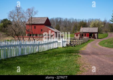 Idyllic colonial Pennsylvania scene with a red wooden barn, and log cabin in the bvcakground and a long path with a white picket fence. Idyllic villag Stock Photo