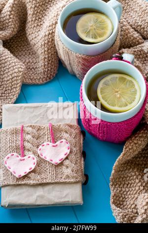 Two blue cup of tea in knitted sweater with hearts felt standing next to the old notebook and blanket knitted on blue background Stock Photo