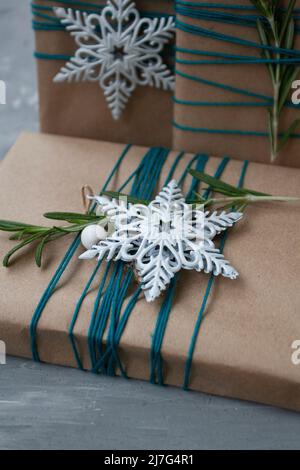 Three Christmas gifts in craft paper wrapped in blue thread, with white snowflakes and rosemary branches on a gray concrete background. There is a pla Stock Photo