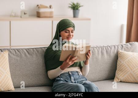 Stay home hobbies. Beautiful young Arab woman in hijab reading book, sitting on couch in living room, copy space Stock Photo