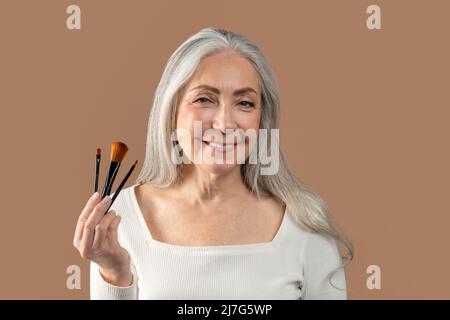 Portrait of beautiful mature woman holding set of different cosmetic brushes over brown studio background Stock Photo