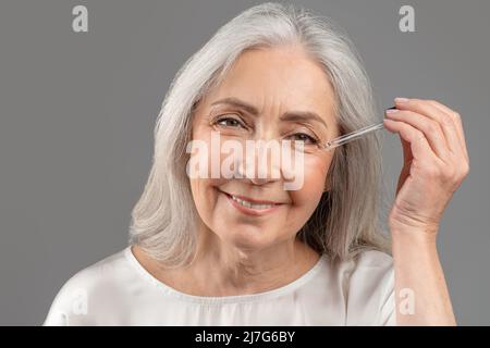 Rejuvenating cosmetics. Beautiful senior woman applying anti-aging serum onto her face over grey studio background Stock Photo
