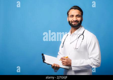 Indian doctor in white medical gown with stethoscope on shoulders taking notes standing with clipboard isolated on blue, physician therapist writing down treatment plan, writes a prescription Stock Photo