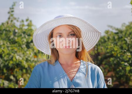Beautiful Caucasian girl in a blue dress and hat among the vineyards in summer Stock Photo