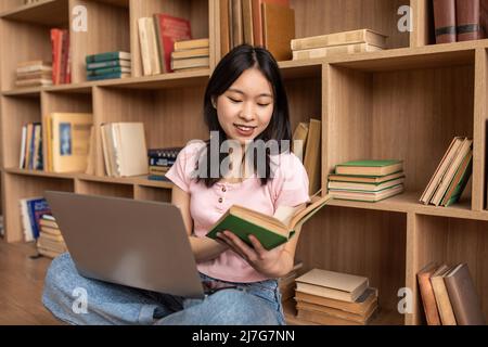 Young korean lady reading book, sitting on floor with laptop pc and study materials at home, copy space Stock Photo