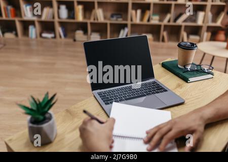 Online class concept. Male student writing in notebook, taking notes and using laptop with black screen for mockup Stock Photo