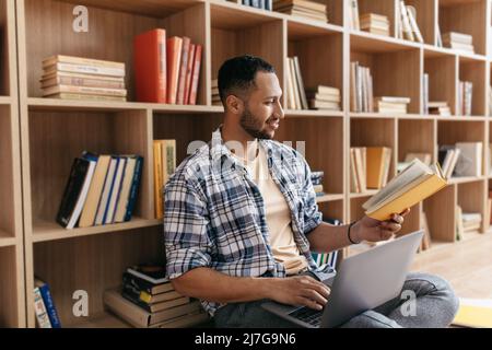 Arab man learning online, reading book and using laptop computer, sitting on floor an leaning on book shelf at home Stock Photo