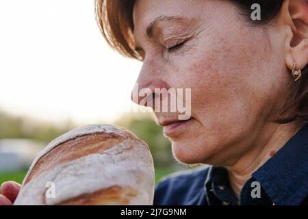 Mature woman baker smelling round loaf of fresh bread at sunset. Stock Photo