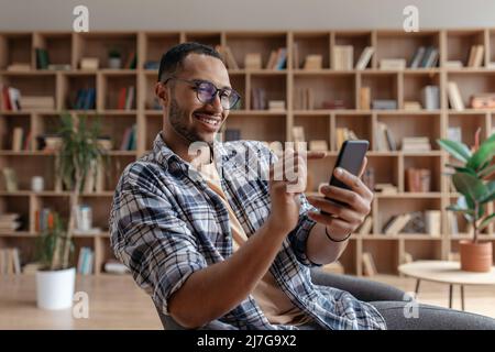 Happy arab young man sitting in chair with cellphone, chatting with friends, resting at home interior, free space Stock Photo