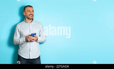 Middle Aged Man Holding Cellphone Looking Aside Over Blue Background Stock Photo
