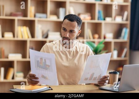 Focused arab man looking through documents, checking reports while having online meeting on laptop, sitting at home Stock Photo