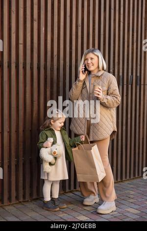 happy woman with paper cup talking on smartphone near daughter with teddy bear and shopping bag Stock Photo