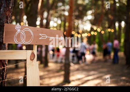 Wooden arrow sign with wedding rings on ceremony venue. Wedding party banner post . Direction info banner for guests in pine forest outdoors. Rustic Stock Photo