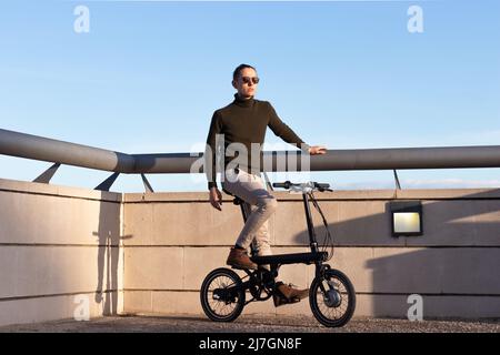 Young man riding an electric bike as a sustainable urban transportation to avoid pollution in the city. Under blue sky with copy space Stock Photo