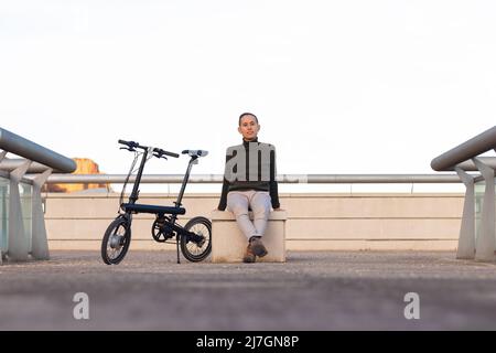Young man sitting on a park bench with his battery bike on the side looking at the camera smiling. with a lot of copy space around Stock Photo