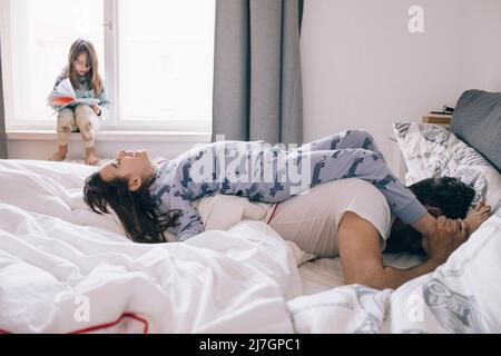 Playful father and daughter lying on bed while girl reading book sitting at window Stock Photo