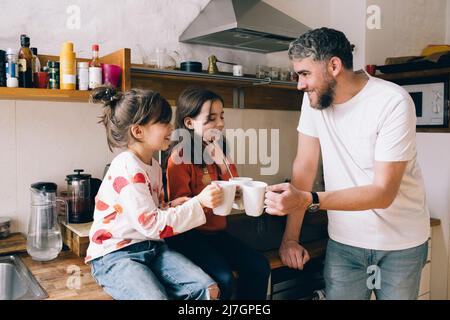 Smiling father toasting mugs with twin daughters sitting on kitchen counter at home Stock Photo
